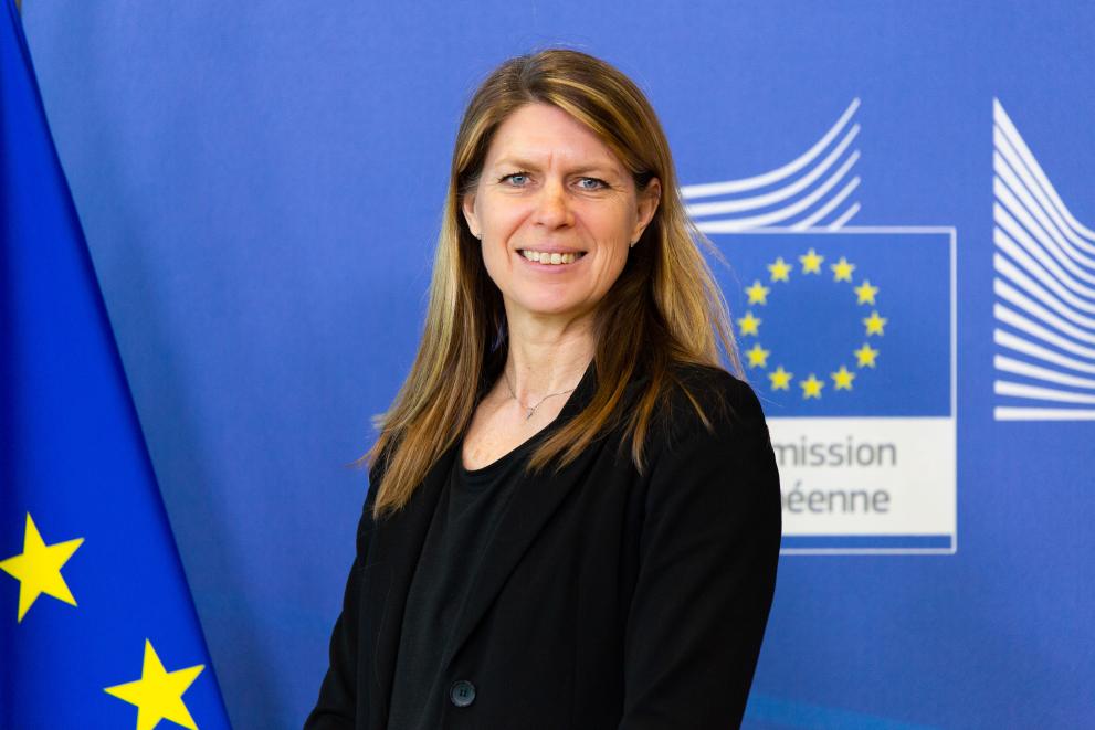 Annika Wäppling Korzinek står bredvid en EU-flagga med kommissionens logo i bakgrunden