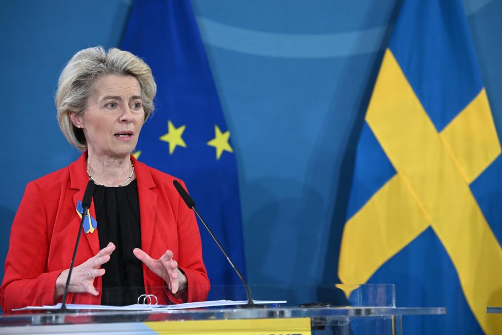 Ursula von der Leyen in red and black speaking at a podium with small hand gestures and an EU flag next to a Swedish flag behind her